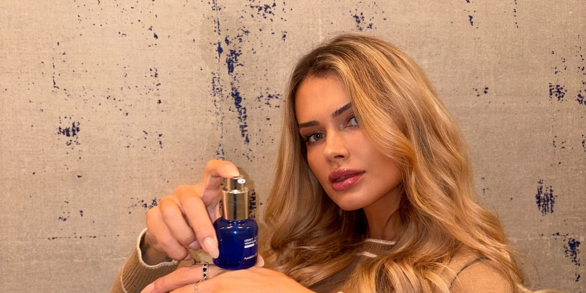 A woman in a camel knit top holds a cobalt Legacy Youth Elixir pump bottle against a textured wall flecked with blue.