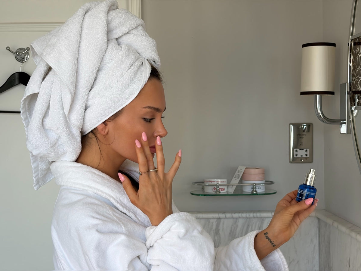 Aurabiom founder applying Legacy Youth Elixir serum to her cheek in a bathroom, holding the blue bottle